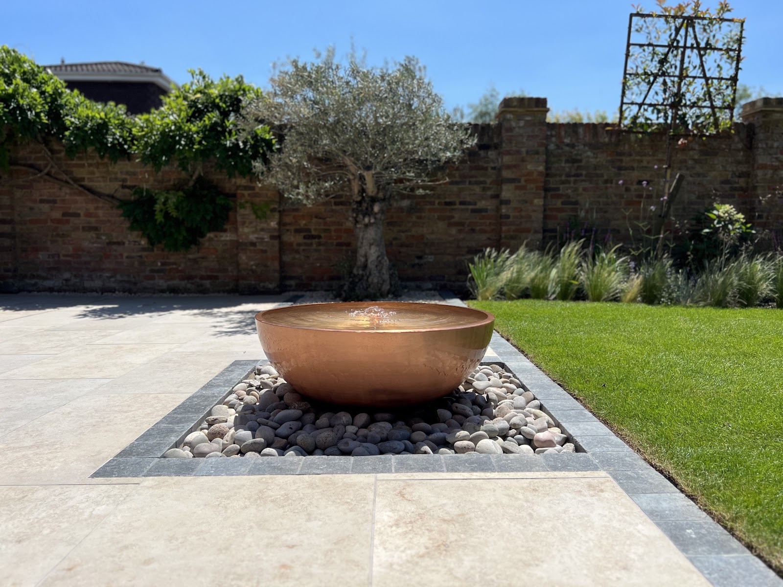 A polished copper bowl water feature set into pebbles and a travertine terrace, framed by a mature olive tree against a period brick garden wall.
