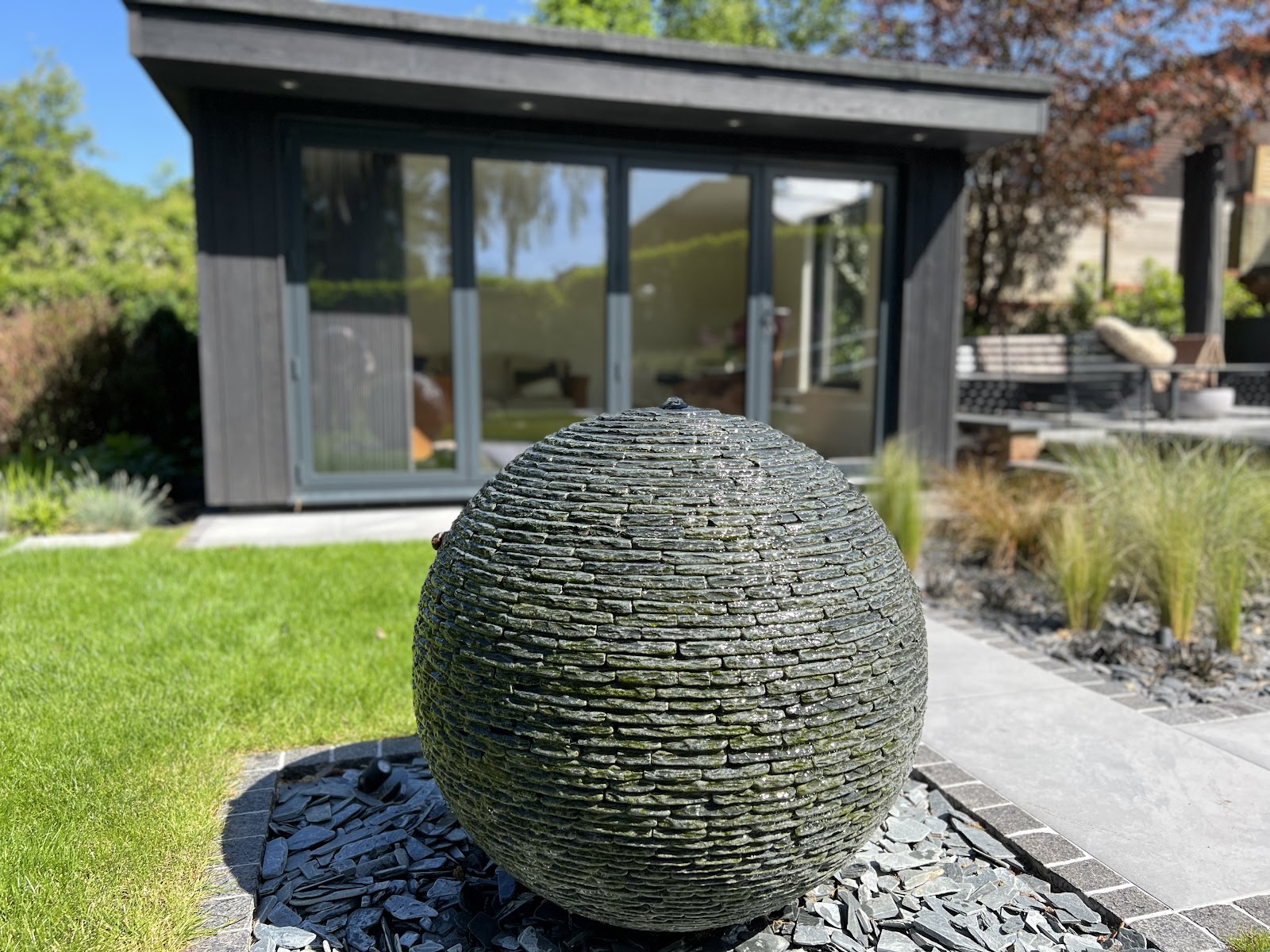 A tall slate-clad sphere water feature on a bed of slate paddlestones, set on a crisp lawn in front of a contemporary garden room with ornamental grasses alongside.